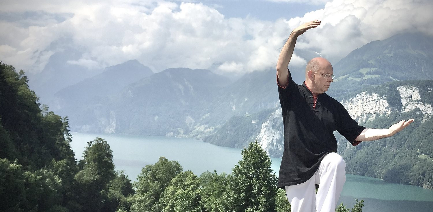 Horst-Rainer Rust übt Qi Gong hoch über einem Bergsee, ein Arm über dem Kopf, der andere zur Seite geöffnet