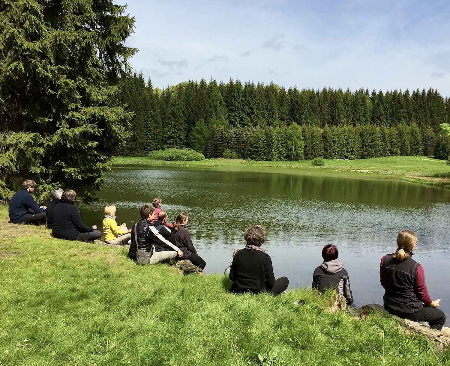 Qi-Gong-Gruppe sitzt zur Meditation am Ufer eines stillen Waldsees
